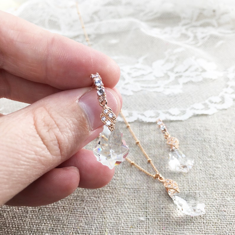 May include: A rose gold jewellery set comprising a necklace and earrings. The necklace and earrings feature clear crystal pendants and are embellished with small, sparkling crystals. The jewellery is set against a soft, neutral backdrop.