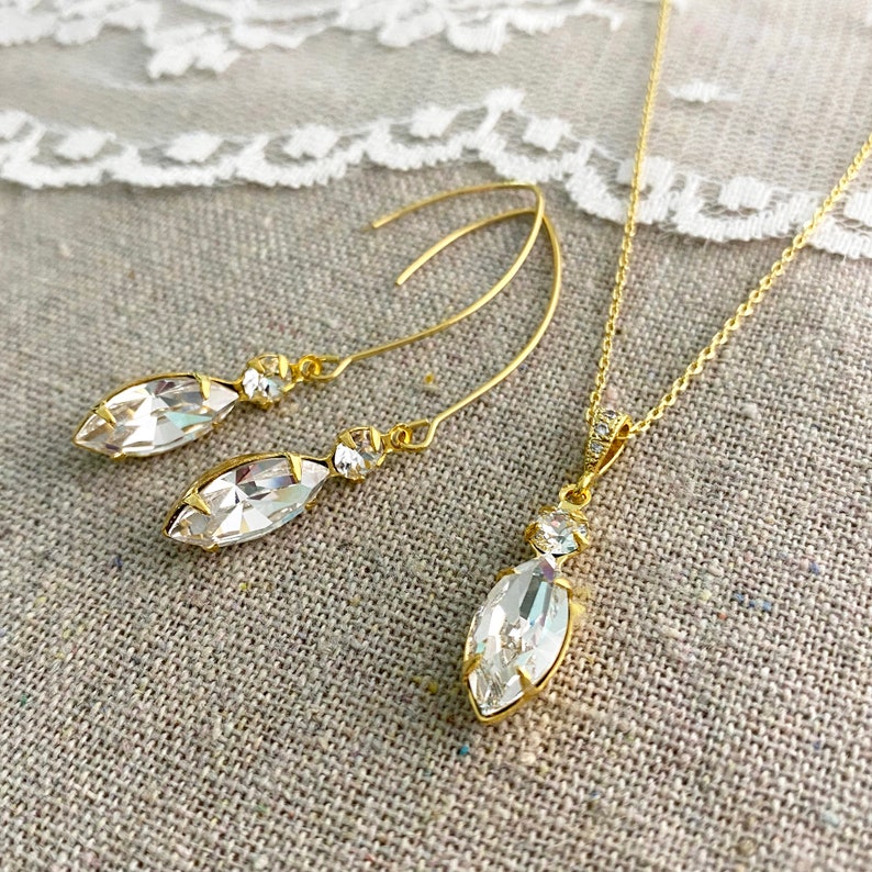 May include: A gold-toned jewelry set featuring a pair of drop earrings and a pendant necklace. Each piece showcases a marquise-cut clear crystal, accented with smaller round crystals. The earrings have a hook design, and the necklace has a delicate chain.
