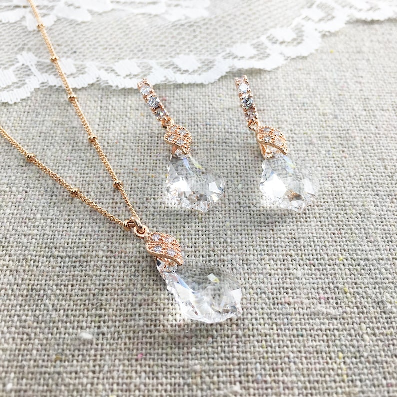 May include: A rose gold jewellery set comprising a necklace and earrings. The necklace features a pendant with a large, clear crystal. The earrings have a similar crystal design. The crystals are teardrop-shaped and faceted, reflecting light. The set is displayed on a textured, neutral-coloured surface.