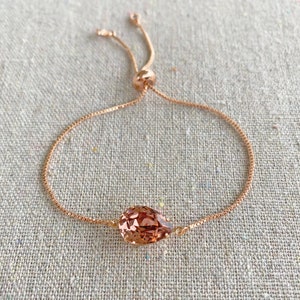 K&ouml;nnte beinhalten: Verstellbares Armband aus Ros&eacute;gold mit einem tropfenf&ouml;rmigen, facettierten Pfirsichkristall. Die feine Kette und der Schiebeverschluss erm&ouml;glichen eine individuelle Anpassung. Das Armband wird auf einer neutralen Leinenoberfl&auml;che gezeigt.