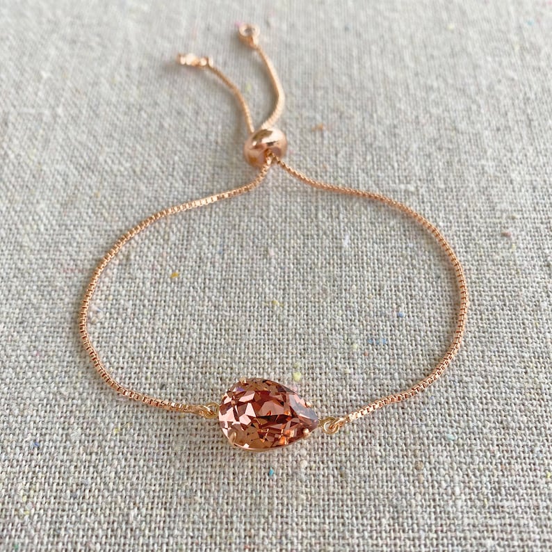 K&ouml;nnte beinhalten: Verstellbares Armband aus Ros&eacute;gold mit einem tropfenf&ouml;rmigen, facettierten Pfirsichkristall. Die feine Kette und der Schiebeverschluss erm&ouml;glichen eine individuelle Anpassung. Das Armband wird auf einer neutralen Leinenoberfl&auml;che gezeigt.