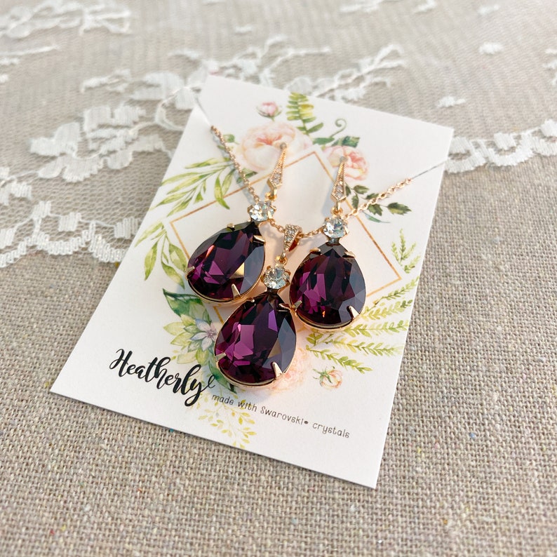 May include: A jewelry set featuring a necklace and earrings with large, teardrop-shaped, deep purple crystals. The crystals are set in a gold-colored metal. The set is displayed on a card with the name "Heatherly" and the text "made with Swarovski crystals."