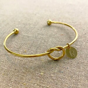 May include: A gold-coloured bangle bracelet featuring a knot design and a small, round charm. The charm is engraved with the letter "B". The bracelet has a simple, elegant design.