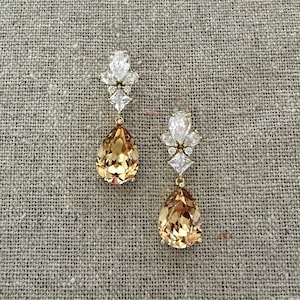May include: A pair of elegant drop earrings. Each earring features a gold-toned setting with a cluster of clear, sparkling crystals at the top. A large, pear-shaped, amber-colored crystal hangs below. The earrings are displayed on a neutral, textured surface.