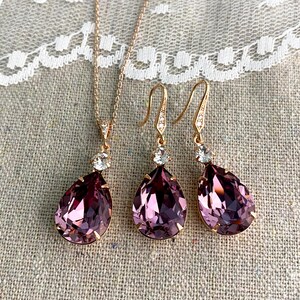 Puede incluir: Un conjunto de joyas de oro rosa que incluye un collar y un par de pendientes. El collar tiene un colgante en forma de l&aacute;grima, de color p&uacute;rpura oscuro, con un acento de cristal transparente. Los pendientes reflejan el dise&ntilde;o del colgante. El conjunto se muestra sobre un fondo de lino neutro.
