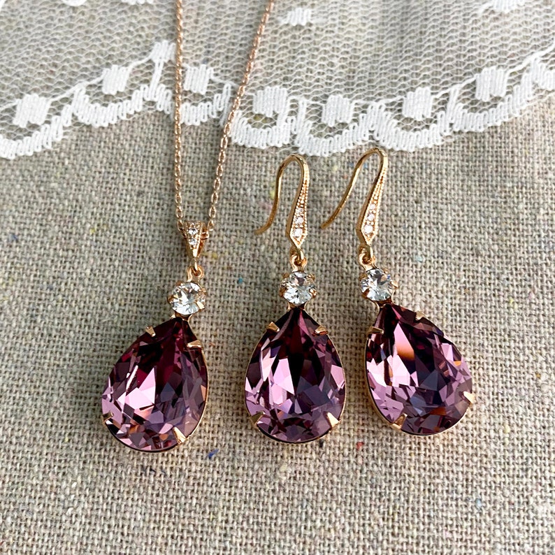 Puede incluir: Un conjunto de joyas de oro rosa que incluye un collar y un par de pendientes. El collar tiene un colgante en forma de l&aacute;grima, de color p&uacute;rpura oscuro, con un acento de cristal transparente. Los pendientes reflejan el dise&ntilde;o del colgante. El conjunto se muestra sobre un fondo de lino neutro.