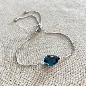 May include: A silver-toned adjustable bracelet featuring a pear-shaped, deep blue gemstone. The bracelet has a delicate chain and a sliding bead for size adjustment. The gemstone is set in a silver bezel.