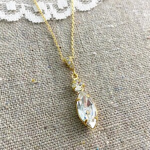 May include: A gold-colored necklace with a clear, marquise-cut pendant. The pendant is suspended from a delicate chain and features a smaller, round, clear stone above the marquise. The necklace is displayed on a textured, neutral-colored surface.