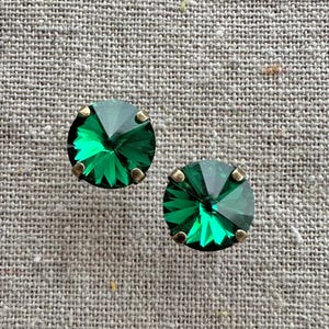 May include: A pair of emerald green stud earrings. Each earring features a faceted, round-cut green gemstone set in a gold-toned metal setting. The earrings are displayed on a textured, neutral-colored surface.