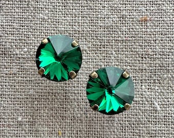 May include: A pair of emerald green stud earrings. Each earring features a faceted, round-cut green gemstone set in a gold-toned metal setting. The earrings are displayed on a textured, neutral-colored surface.