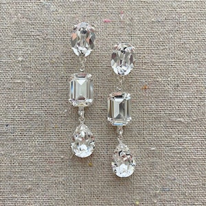 May include: A pair of elegant, clear crystal drop earrings. Each earring features an oval-shaped top stone, a rectangular center stone, and a pear-shaped bottom stone, all connected by silver-toned links. The earrings are displayed against a neutral background.