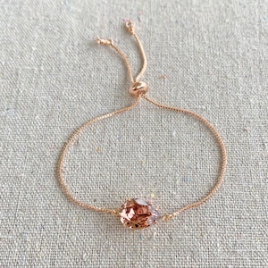 K&ouml;nnte beinhalten: Verstellbares Armband aus Ros&eacute;gold mit einem tropfenf&ouml;rmigen, facettierten Pfirsichkristall. Die feine Kette hat eine verschiebbare Perle zur Gr&ouml;&szlig;enanpassung. Das Armband wird auf einer neutralen Leinenoberfl&auml;che gezeigt.