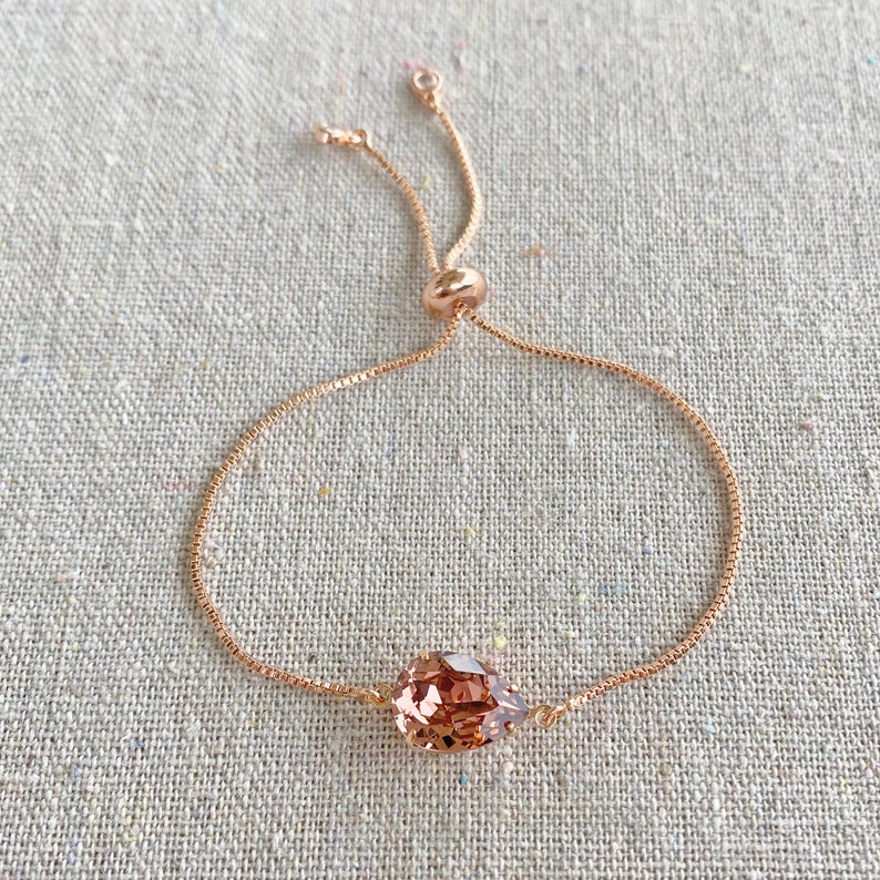 K&ouml;nnte beinhalten: Verstellbares Armband aus Ros&eacute;gold mit einem tropfenf&ouml;rmigen, facettierten Pfirsichkristall. Die feine Kette hat eine verschiebbare Perle zur Gr&ouml;&szlig;enanpassung. Das Armband wird auf einer neutralen Leinenoberfl&auml;che gezeigt.