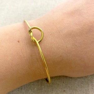 May include: A gold-coloured bangle bracelet featuring a knot design. The bracelet is slender and delicate, with a simple, elegant style. The knot is the focal point, adding a unique touch to the jewellery.