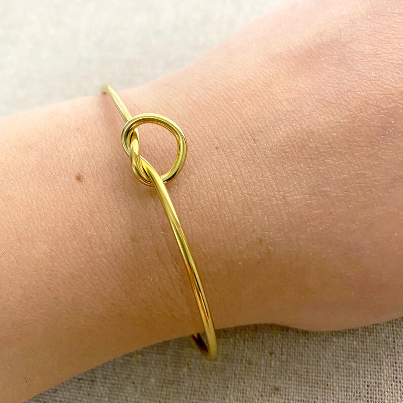 May include: A gold-coloured bangle bracelet featuring a knot design. The bracelet is slender and delicate, with a simple, elegant style. The knot is the focal point, adding a unique touch to the jewellery.
