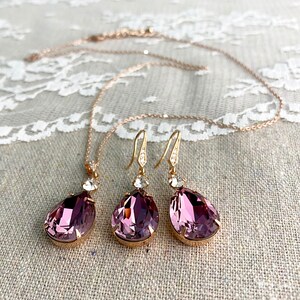 Puede incluir: Un conjunto de joyas que incluye un collar y pendientes. El conjunto presenta un collar de cadena de oro rosa con un colgante en forma de l&aacute;grima de color p&uacute;rpura oscuro. Los pendientes tienen un dise&ntilde;o similar, con una piedra p&uacute;rpura en forma de l&aacute;grima y ganchos dorados.