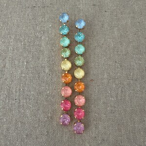 May include: A pair of long drop earrings featuring a vertical row of ten round, faceted crystals in a gradient of colors ranging from light blue at the top to purple at the bottom, all set in a thin gold-toned metal frame with small prongs.