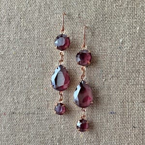 May include: A pair of drop earrings featuring rose gold-toned metal settings and faceted, deep burgundy-colored stones. Each earring consists of a round stone at the top, a larger pear-shaped stone in the center, and a small round stone at the base.