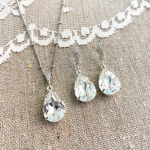 May include: A silver necklace with a teardrop-shaped pendant and matching teardrop earrings. The jewelry set features clear, faceted crystals, and is displayed on a neutral-toned fabric with a lace trim.