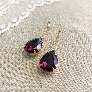 May include: A pair of gold-toned drop earrings with large, faceted, deep purple teardrop-shaped crystals. Each earring has a small, clear crystal accent and a decorative hook. The earrings are displayed on a neutral linen surface.