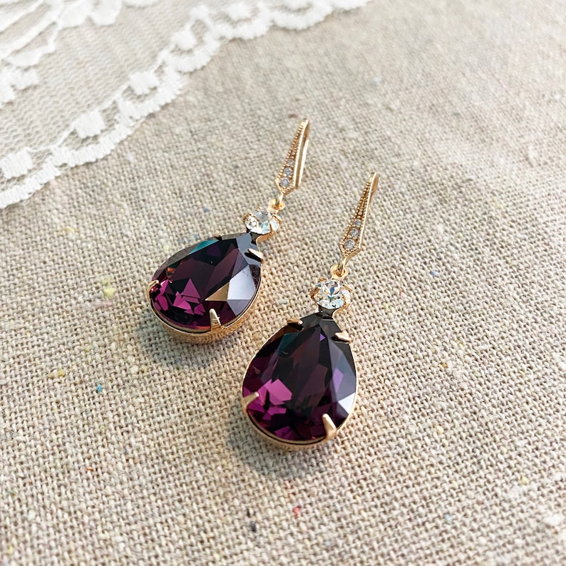May include: A pair of gold-toned drop earrings with large, faceted, deep purple teardrop-shaped crystals. Each earring has a small, clear crystal accent and a decorative hook. The earrings are displayed on a neutral linen surface.