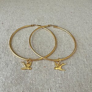 May include: A pair of gold-colored hoop earrings. Each earring has a small gold bird charm dangling from the bottom. The earrings are displayed on a neutral-colored, textured surface.