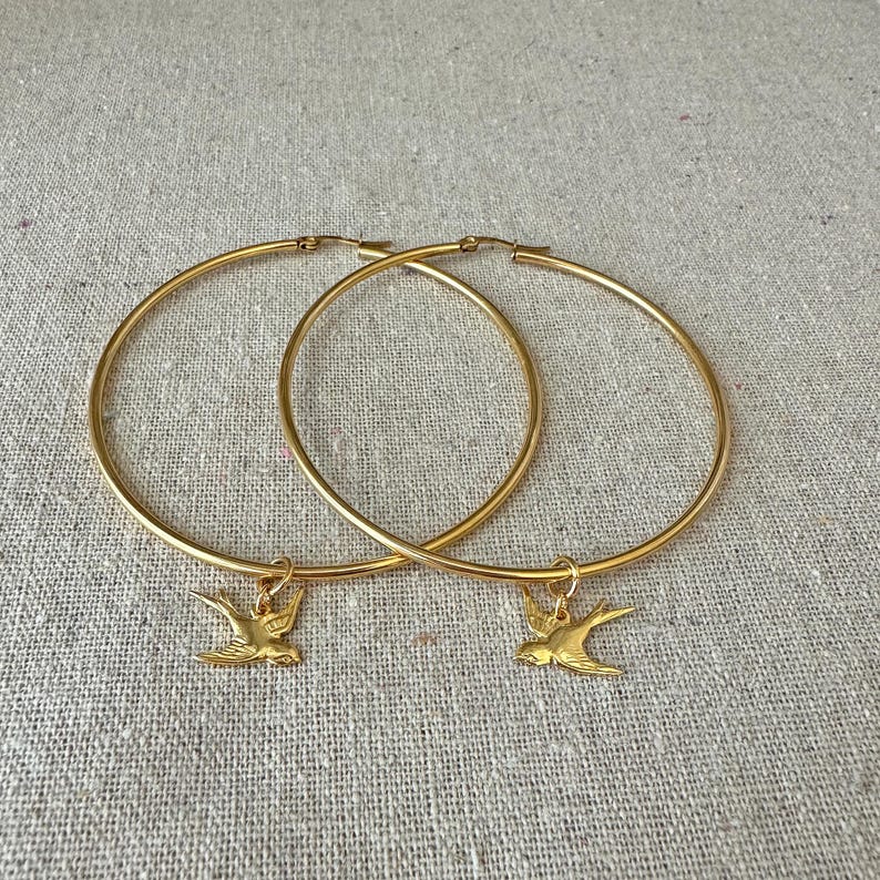 May include: A pair of gold-colored hoop earrings. Each earring has a small gold bird charm dangling from the bottom. The earrings are displayed on a neutral-colored, textured surface.