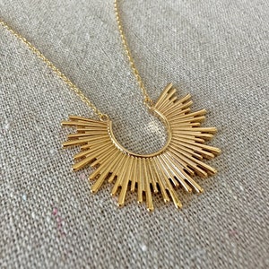 May include: A gold-colored necklace featuring a sunburst pendant. The pendant has a half-circle design with radiating lines, creating a sun-like appearance. The necklace chain is delicate and also gold-toned. The jewelry is displayed on a neutral, textured surface.