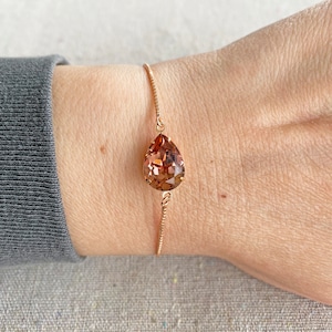 K&ouml;nnte beinhalten: Ein ros&eacute;goldenes Armband mit einem gro&szlig;en, tropfenf&ouml;rmigen, facettierten Kristall in Braun- und Pfirsicht&ouml;nen. Der Kristall h&auml;ngt an einer feinen Kette. Das Armband wird am Handgelenk gezeigt.