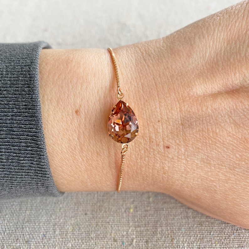 K&ouml;nnte beinhalten: Ein ros&eacute;goldenes Armband mit einem gro&szlig;en, tropfenf&ouml;rmigen, facettierten Kristall in Braun- und Pfirsicht&ouml;nen. Der Kristall h&auml;ngt an einer feinen Kette. Das Armband wird am Handgelenk gezeigt.