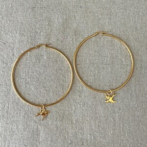 May include: A pair of gold-colored hoop earrings. Each earring features a small, gold-colored bird charm dangling from the bottom. The earrings are displayed on a neutral-colored, textured surface.