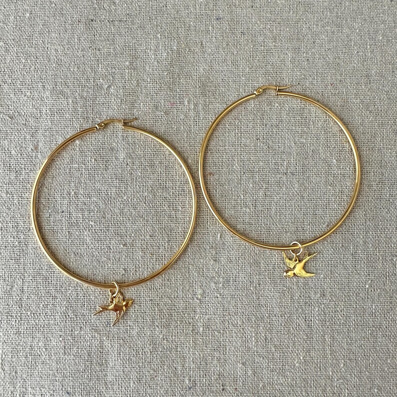 May include: A pair of gold-colored hoop earrings. Each earring features a small, gold-colored bird charm dangling from the bottom. The earrings are displayed on a neutral-colored, textured surface.