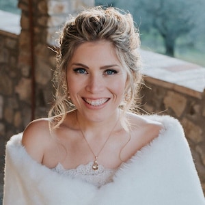 May include: A woman with blonde hair and blue eyes smiles, wearing a white fur shawl and a delicate necklace. The image is a close-up, highlighting the woman's features and the texture of the shawl. The background is blurred, suggesting an outdoor setting.