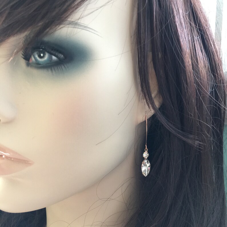 May include: A pair of elegant drop earrings. Each earring features a small, round, clear crystal above a larger, marquise-shaped crystal. The crystals are set on a thin, gold-colored wire. The earrings are shown on a mannequin.