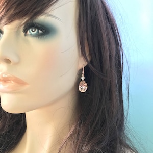 Puede incluir: Pendientes de l&aacute;grima de oro rosa con un acento de cristal transparente. Los pendientes presentan un cristal facetado en forma de pera, de color melocot&oacute;n. Se muestran en un maniqu&iacute; con cabello casta&ntilde;o y maquillaje de ojos azul.