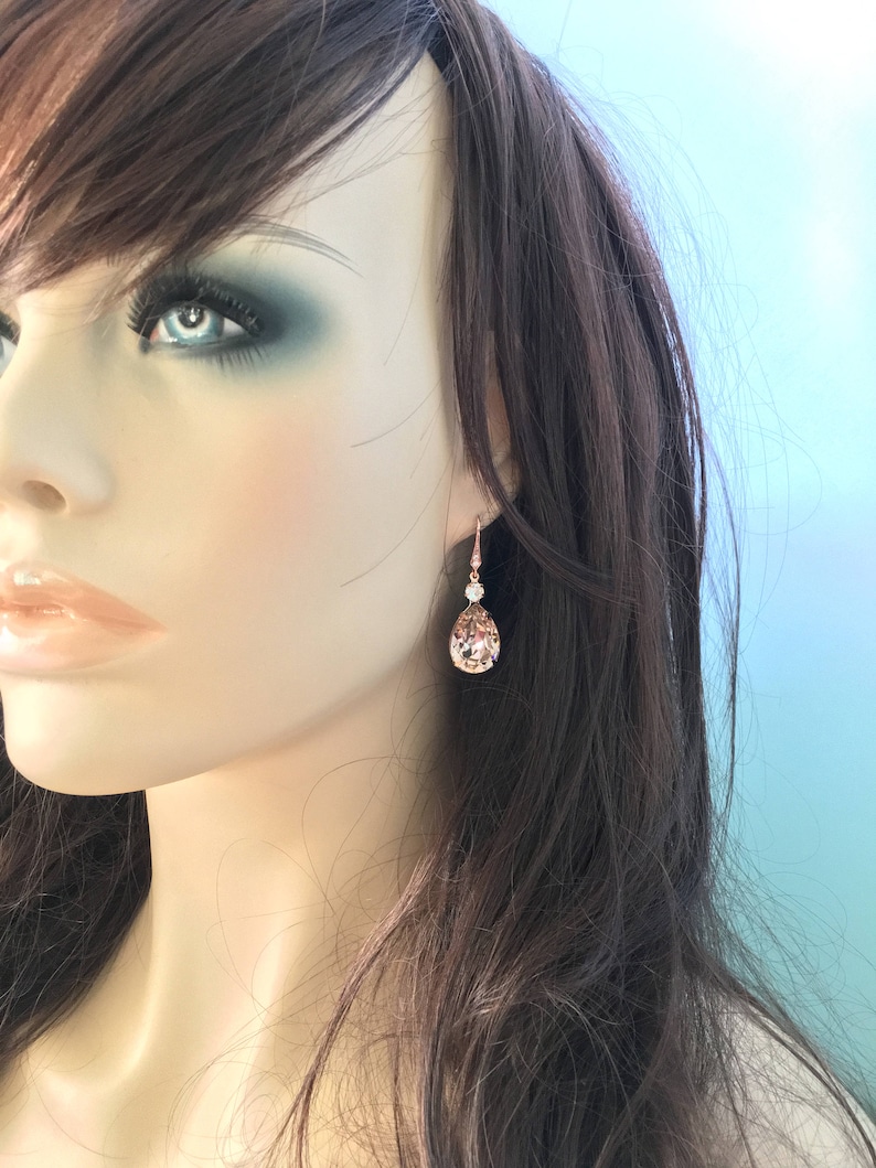 Puede incluir: Pendientes de l&aacute;grima de oro rosa con un acento de cristal transparente. Los pendientes presentan un cristal facetado en forma de pera, de color melocot&oacute;n. Se muestran en un maniqu&iacute; con cabello casta&ntilde;o y maquillaje de ojos azul.