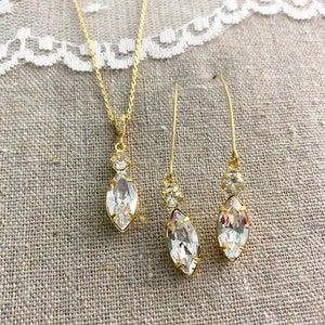 May include: A gold-toned jewelry set featuring a necklace and a pair of earrings. The necklace has a pendant with a large, clear, marquise-cut crystal. The earrings have a similar design, with long, slender hooks and marquise-cut crystals. The set is displayed on a neutral linen surface.
