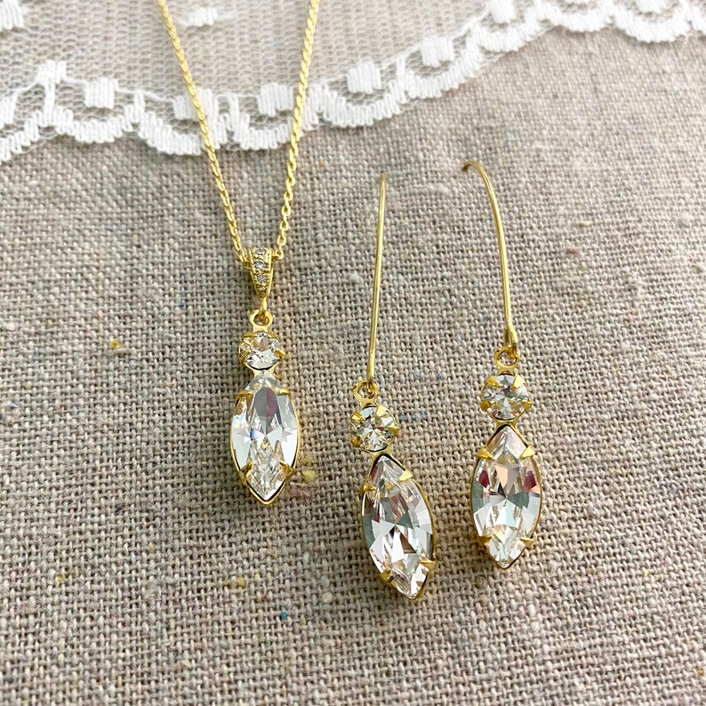 May include: A gold-toned jewelry set featuring a necklace and a pair of earrings. The necklace has a pendant with a large, clear, marquise-cut crystal. The earrings have a similar design, with long, slender hooks and marquise-cut crystals. The set is displayed on a neutral linen surface.