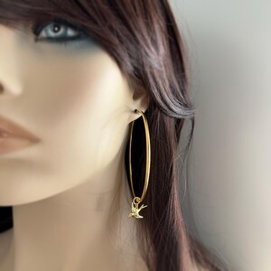 May include: Gold-colored hoop earrings with a small bird charm. The earrings are long and oval-shaped, with a simple, elegant design. The bird charm is a delicate, detailed silhouette.