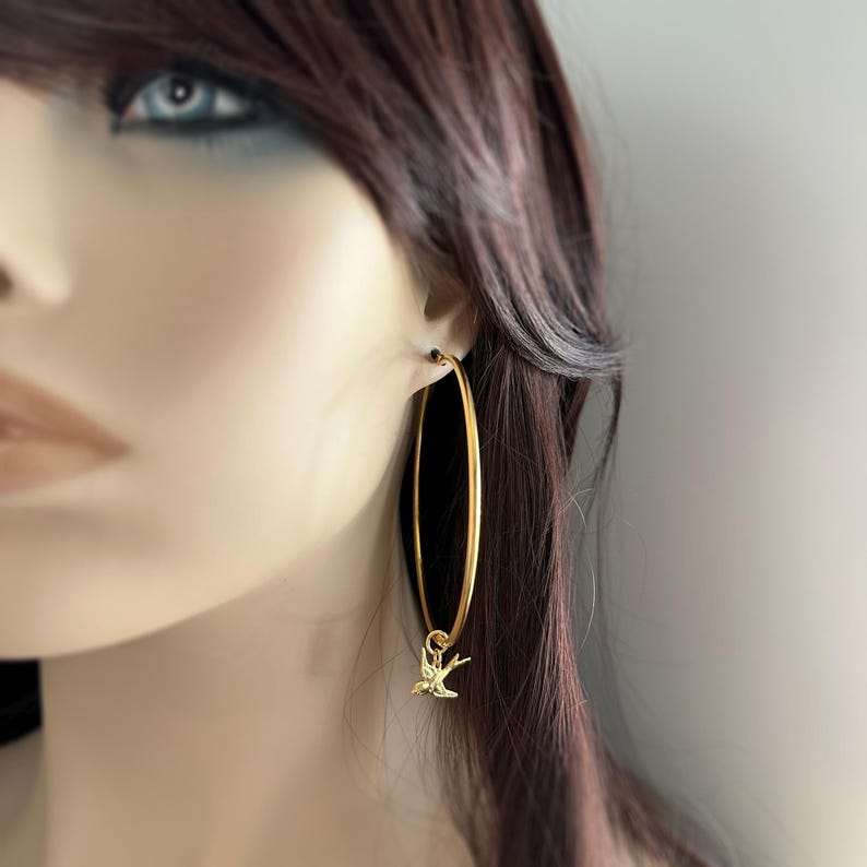 May include: Gold-colored hoop earrings with a small bird charm. The earrings are long and oval-shaped, with a simple, elegant design. The bird charm is a delicate, detailed silhouette.