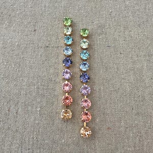 May include: A pair of gold-toned earrings, each featuring a vertical line of ten colorful, round gemstones. The gemstones transition in color from light green to peach. The earrings are displayed on a neutral, textured background.