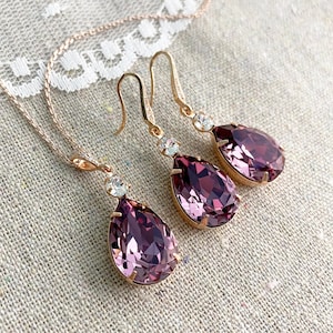 Puede incluir: Un conjunto de joyas de oro rosa con un collar y un par de pendientes. Cada pieza tiene un colgante de cristal p&uacute;rpura grande en forma de l&aacute;grima y un acento de cristal transparente redondo m&aacute;s peque&ntilde;o. Las joyas se exhiben sobre una tela de color neutro.