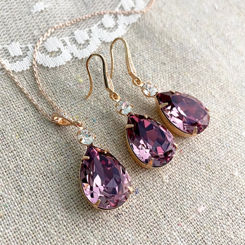 Puede incluir: Un conjunto de joyas de oro rosa con un collar y un par de pendientes. Cada pieza tiene un colgante de cristal p&uacute;rpura grande en forma de l&aacute;grima y un acento de cristal transparente redondo m&aacute;s peque&ntilde;o. Las joyas se exhiben sobre una tela de color neutro.