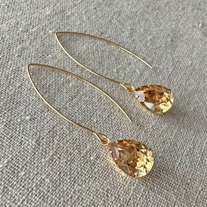 May include: A pair of gold-colored drop earrings. Each earring features a long, curved wire with a teardrop-shaped, amber-colored crystal pendant. The earrings are displayed on a neutral-colored, textured surface.
