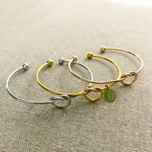 May include: Four open cuff bracelets in silver, gold, and rose gold tones. Two bracelets feature a knot design, and one has a small round charm with an initial. The bracelets are displayed on a neutral linen surface.