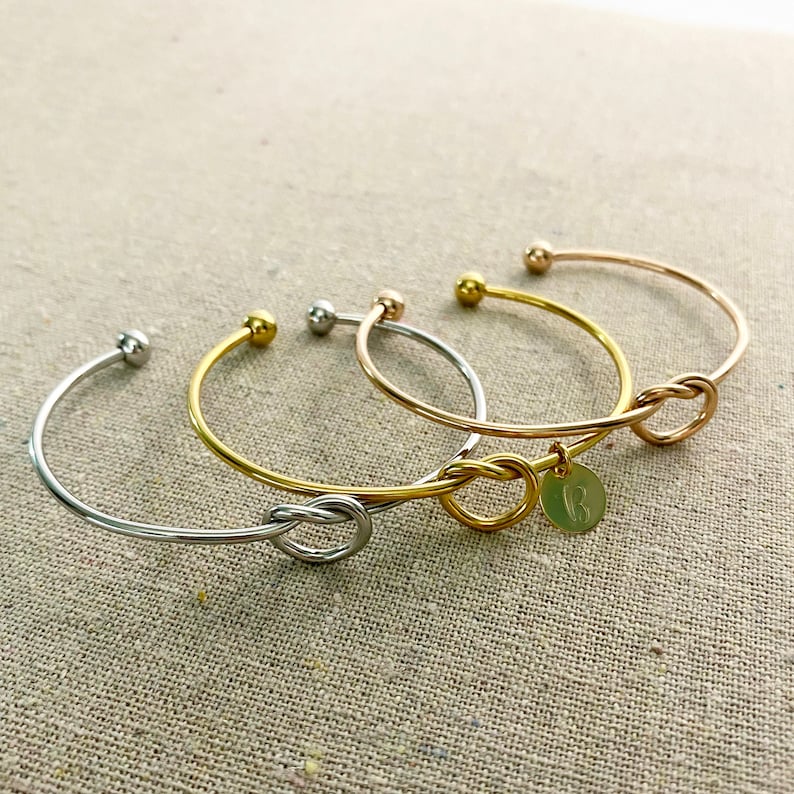 May include: Four open cuff bracelets in silver, gold, and rose gold tones. Two bracelets feature a knot design, and one has a small round charm with an initial. The bracelets are displayed on a neutral linen surface.