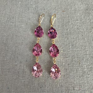 May include: A pair of dangle earrings with three pink teardrop-shaped crystals. The crystals are connected by gold-colored chains and have gold-colored ear wires. The earrings are displayed on a textured, neutral-colored surface.