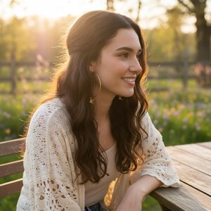 May include: A woman wearing large gold hoop earrings with a small charm, a cream-colored open-knit cardigan, and a white top. She is smiling, with long brown hair, and is sitting at a wooden table outdoors.