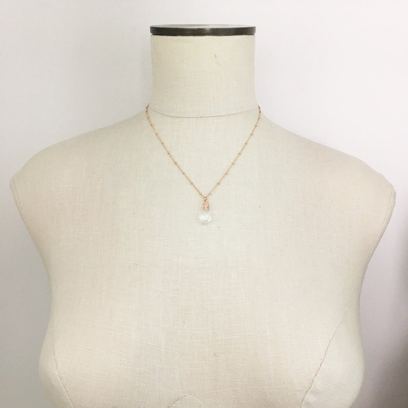 May include: A delicate rose gold necklace with a small, clear crystal pendant. The necklace features a fine chain with small, evenly spaced beads. The crystal pendant is faceted, catching the light. The necklace is displayed on a mannequin.