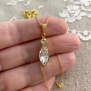 May include: A gold-colored necklace with a large, clear marquise-cut crystal pendant. The pendant is suspended from a delicate chain and features a smaller round crystal above it. The necklace is held against a neutral background.